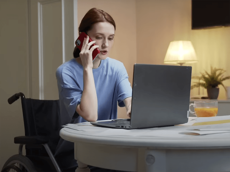Woman in wheelchair using laptop and phone at table with tea and lamp implementing Syntheia's customer support