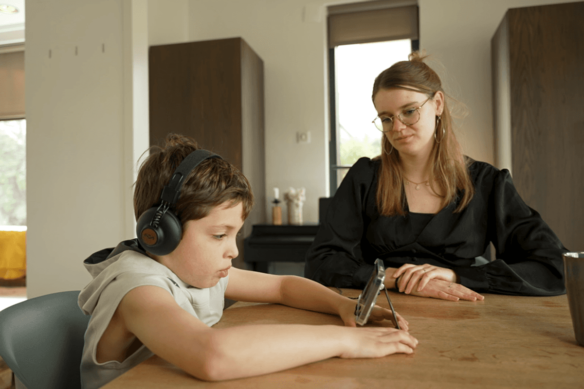 Woman watches child wearing headphones use tablet.