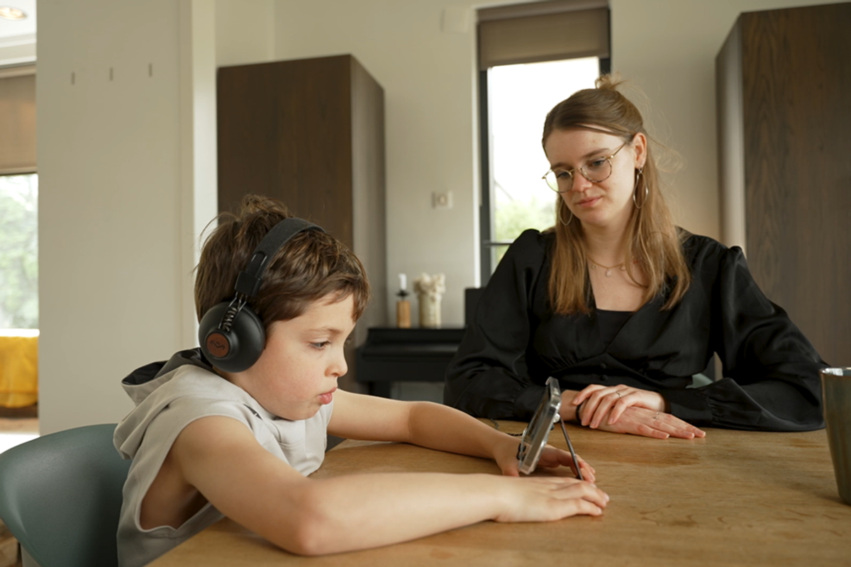 Woman watches child wearing headphones use tablet.