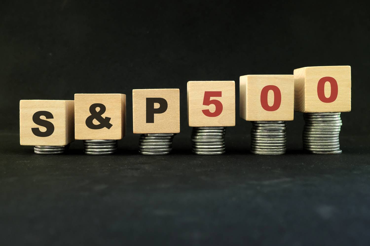 Wooden blocks spelling "S&P 500" are stacked on piles of coins against a black background.
