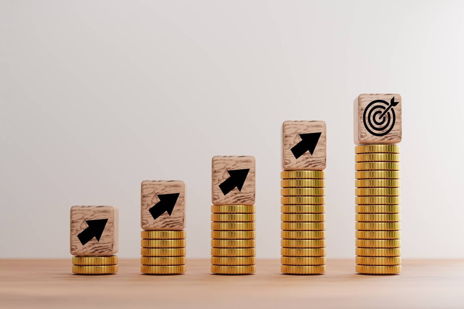 Wooden blocks with arrows and target symbols sit atop ascending stacks of gold coins on a wooden surface.
