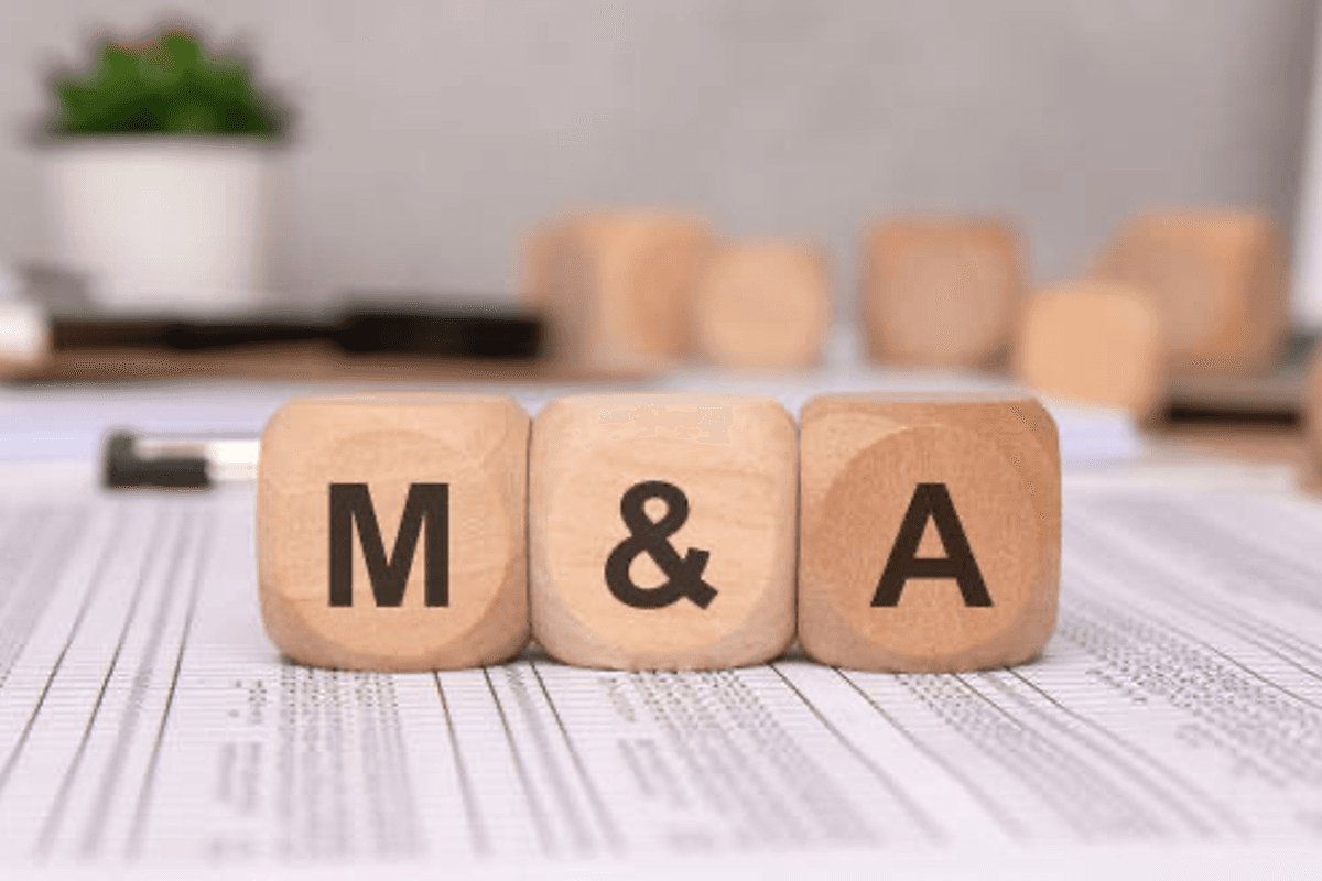 Wooden blocks with "M&A" on a table over documents, plant in background.