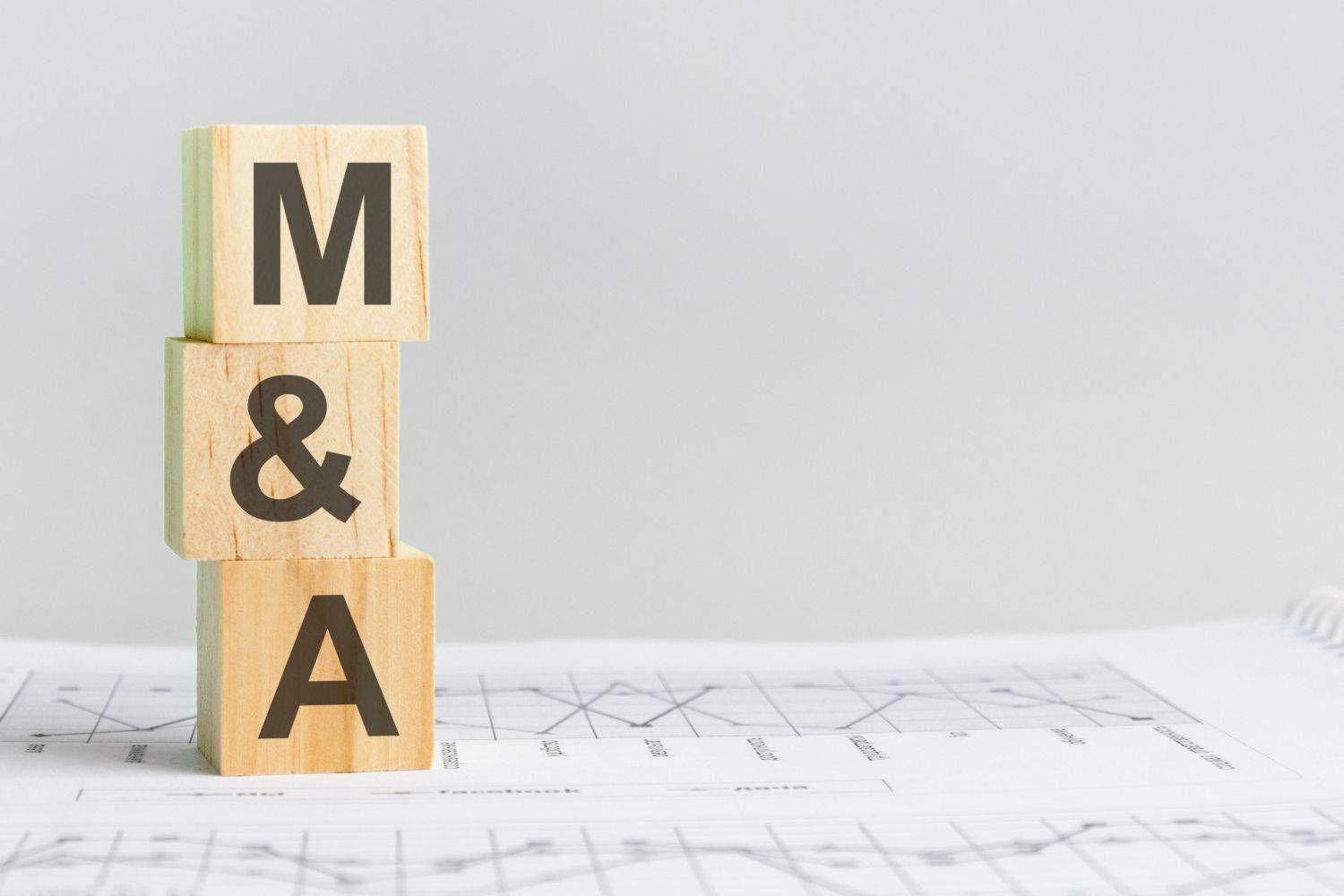 Wooden blocks with "M & A" stand vertically on white papers with charts, against a gray background.