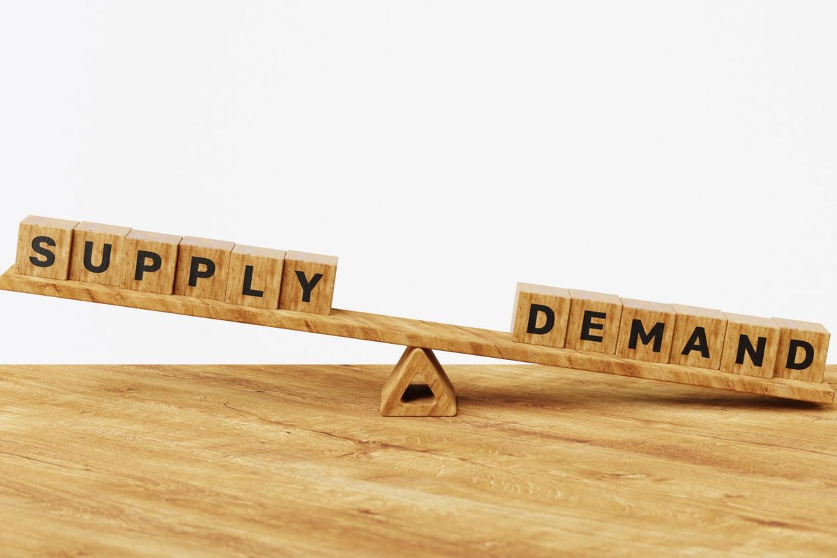 Wooden seesaw with "supply" and "demand" blocks, tilting towards demand.