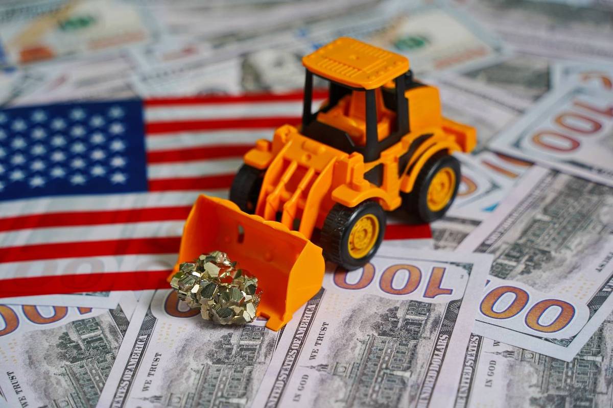 Yellow toy bulldozer scooping gold nugget on US$100 bills and American flag.