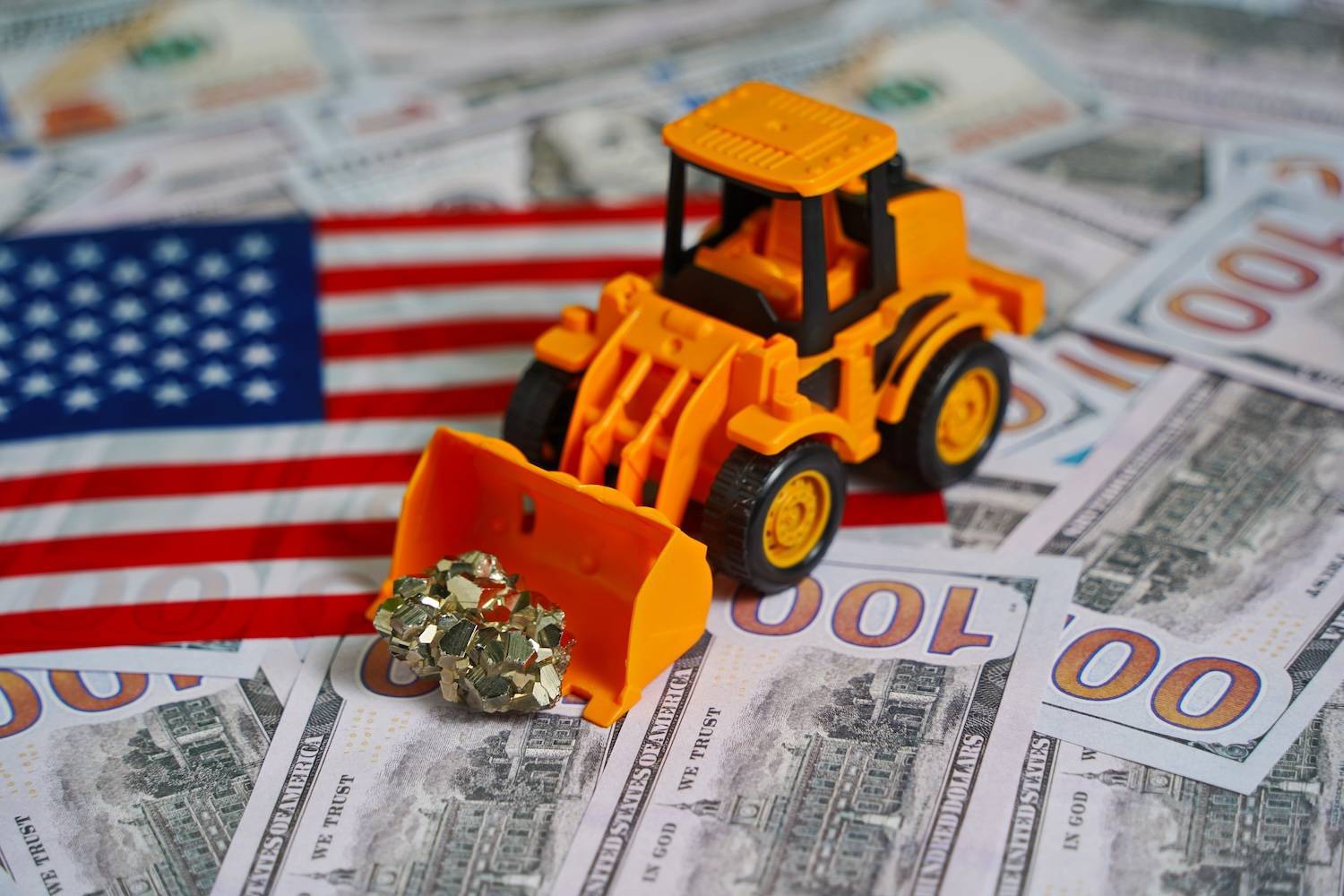 Yellow toy bulldozer scooping gold nugget on US$100 bills and American flag.